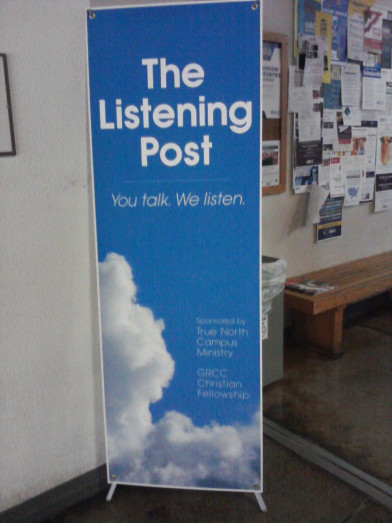 Picture of a banner on a college campus. The vertical banner has blue skies and clouds in the background with a white border. The words on the banner reads "The Listening Post. You Talk. We Listen."