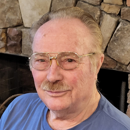 White male with gray and brown muchstach and hair. He is wearing glasses. Looking at the camera smiling. He is wearing a light blue shirt. The background is stone backdrop.